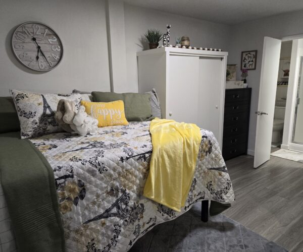 Cozy bedroom with a twin bed, floral quilt, green throw, yellow accent pillow, and a yellow blanket; a white wardrobe nearby.
