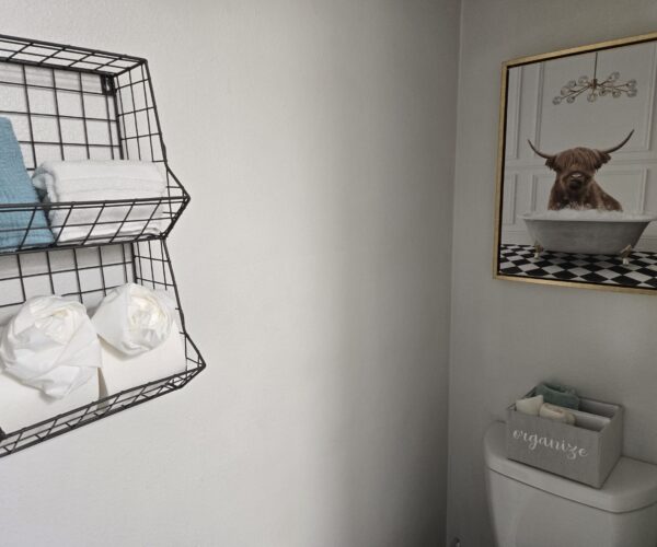 Bathroom corner with a wire wall shelf holding blue and white towels and toilet paper, plus a framed Highland cow in a bathtub art print on the wall to the right of the toilet.