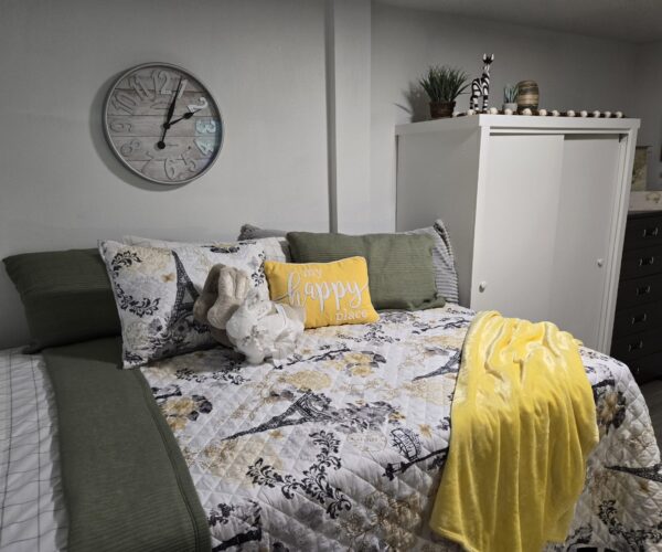 Cozy bedroom with a floral quilt, green pillows, a yellow decorative pillow, and a yellow throw on the bed; a large clock on the wall above.