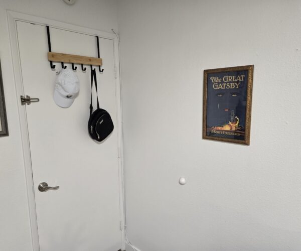 White interior door with a wooden coat rack holding a white cap and black bag, and a framed The Great Gatsby poster on the adjacent wall.