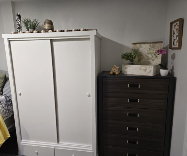White wardrobe beside a dark wooden dresser in a cozy bedroom; decor on top includes a map box, plants, and a teddy bear.