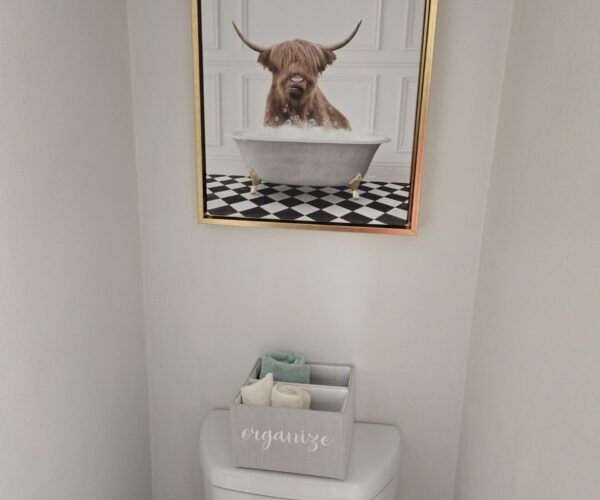 Framed photograph of a Highland cow in a bathtub on a checkered floor, mounted above a white toilet.