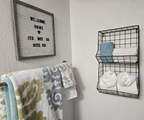 Framed letter-board greeting that says 'Welcome Home' with a small heart, mounted on a textured wall above a decorative towel rack with patterned towels and a blue towel on the shelf beside it.