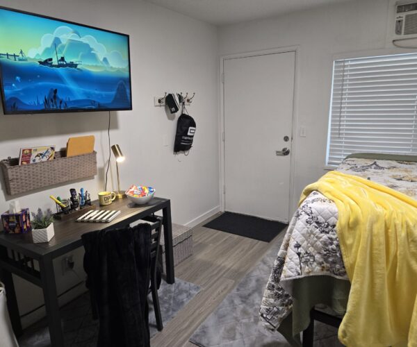 Bedroom with a wall-mounted TV above a dark desk, desk supplies, lamp, and a bowl of snacks; yellow blanket on the bed to the right.