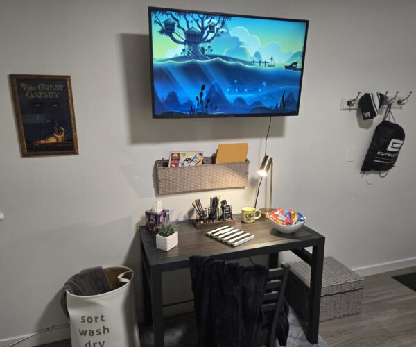 Desk setup under a wall-mounted TV with a basket shelf, lamp, pencils, snacks, and a bowl of candy on a dark wood desk; poster on the left wall and a coat rack with bags on the right.