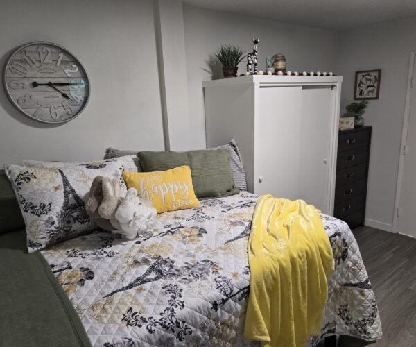 Cozy bedroom with a bed dressed in a patterned quilt, a yellow “my happy place” pillow, a plush bunny, and a yellow throw; white wardrobe and dark chest of drawers nearby; a large wall clock on the left wall.