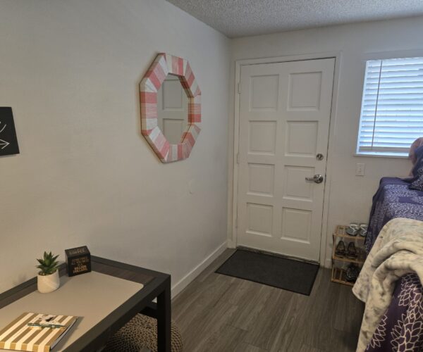 Corner of a bedroom with a pink striped octagonal mirror on the wall, next to a white door, and a window with blinds; bed with purple patterned bedding visible.