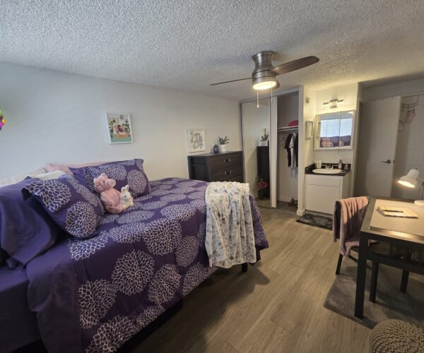 Bedroom scene with purple floral bedding, stuffed animals, and a ceiling fan overhead.