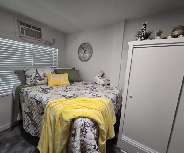 Cozy bedroom with a bed, yellow accents, and a white wardrobe against a light wall. Window blinds and an A/C unit are above the bed; decorative items sit on the wardrobe.