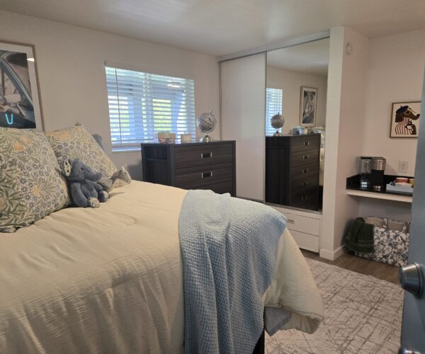 Cozy bedroom with a bed in the foreground, decorative pillows and blue knit blanket, plush toys, and a dark wood dresser against the wall with a mirrored closet nearby.