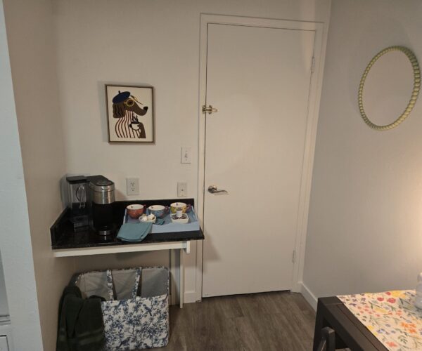 Coffee station on a black countertop with a coffee maker, mugs, and a framed dachshund illustration on the wall.