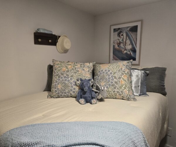 Bed with floral pillows and a blue teddy bear; framed photo of a woman in a car on the beige wall behind.