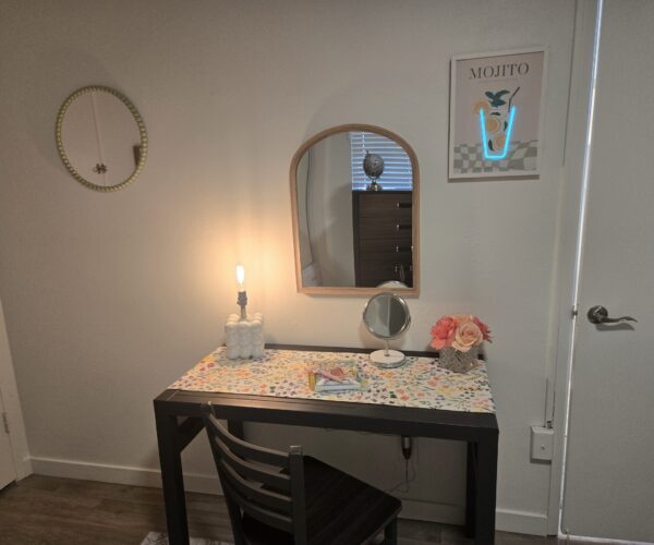 A vanity setup with a table, floral runner, white bead lamp, round wall mirror, and a poster reading 'MOJITO' on the wall to the right, plus a blue neon U-shape.