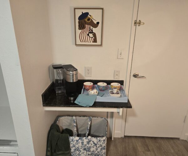 Coffee station on a black countertop with a stainless coffee maker and colorful teacups on a tray; wall art of a cartoon dog hangs above.