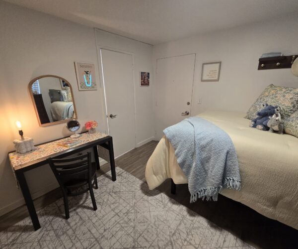 Bedroom scene: bed on the right with blue throw and floral pillows, and a desk with mirror and lamp on the left, creating a cozy space.