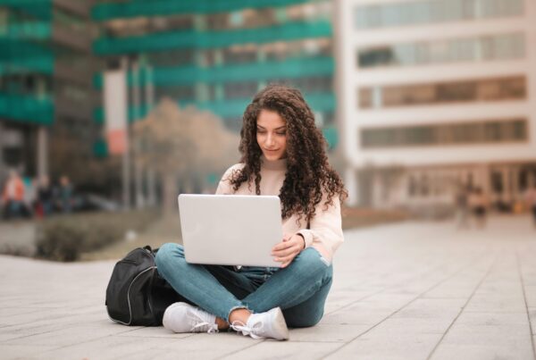 Woman with long curly hair sits cross-legged on a paved plaza, typing on a silver laptop with a black backpack beside her; blurred green and beige city buildings in the background.