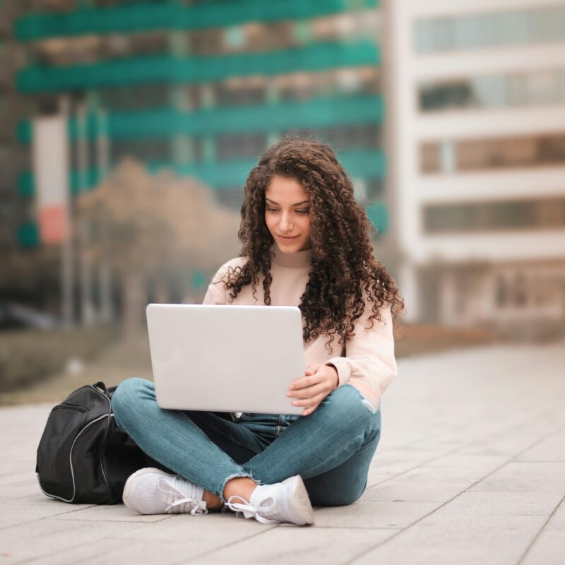 Woman with long curly hair sits cross-legged on a paved plaza, typing on a silver laptop with a black backpack beside her; blurred green and beige city buildings in the background.