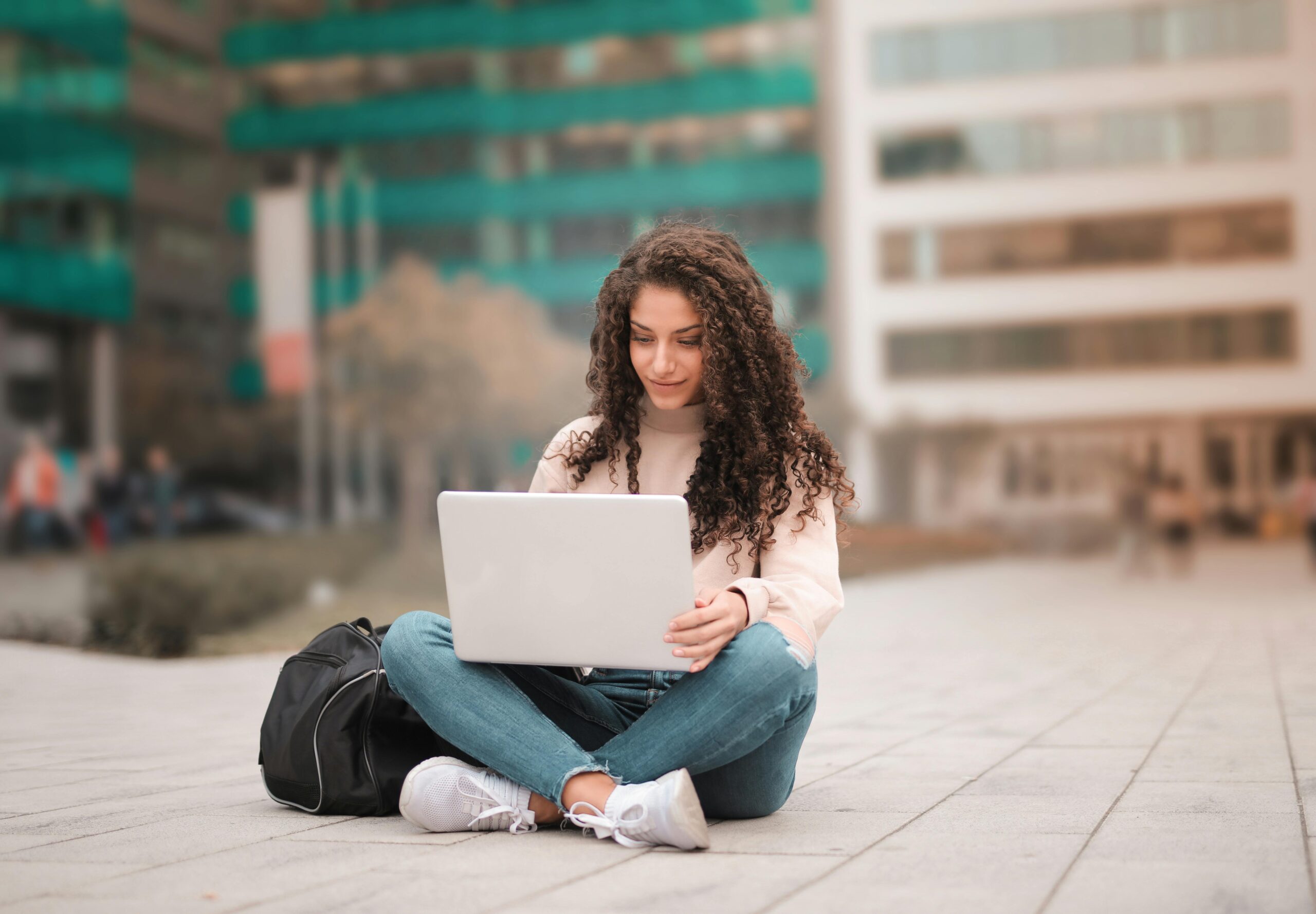 Woman with long curly hair sits cross-legged on a paved plaza, typing on a silver laptop with a black backpack beside her; blurred green and beige city buildings in the background.